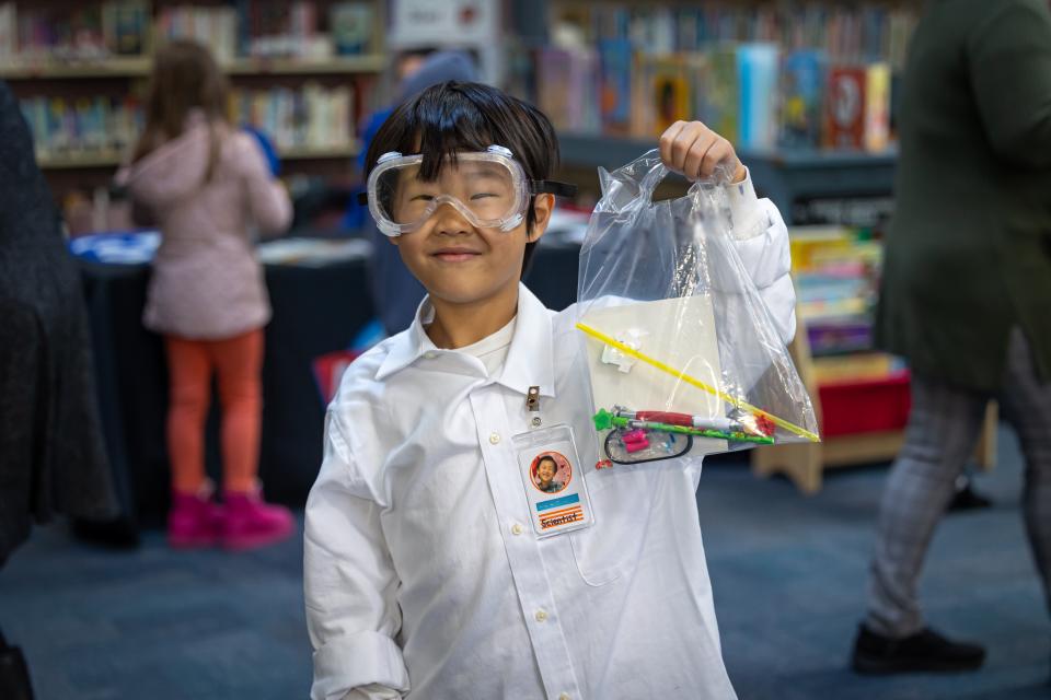 Elementary School student dressed in a lab coat and goggles stands in a library, holding up a plastic bag of school supplies.