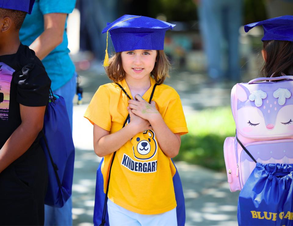 An elementary school student wearing a graduation cap smiling outside. 